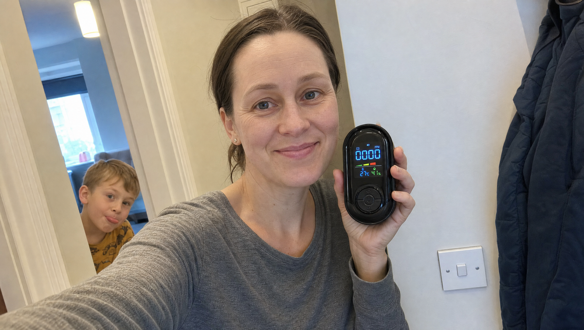 A mother takes a selfie in her hallway holding up her TrueDetect carbon monoxide detector, her young son leaning into the shot from the doorway behind her.